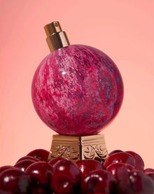 Red marble perfume bottle with gold base surrounded by cherries on a pink background