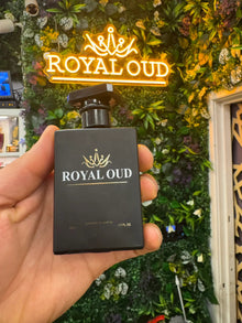 Hand holding Royal Oud perfume bottle with lush indoor plant wall and neon sign background