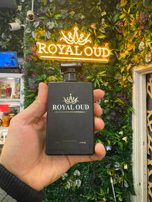 Hand holding Royal Oud perfume bottle in front of a lush green wall with neon Royal Oud sign