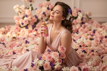 Woman in pink dress holding floral perfume bottle surrounded by roses and violets