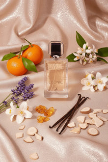 Leonie perfume bottle surrounded by oranges, vanilla, amber, wood, white blossoms, and lavender on a shimmering fabric background.