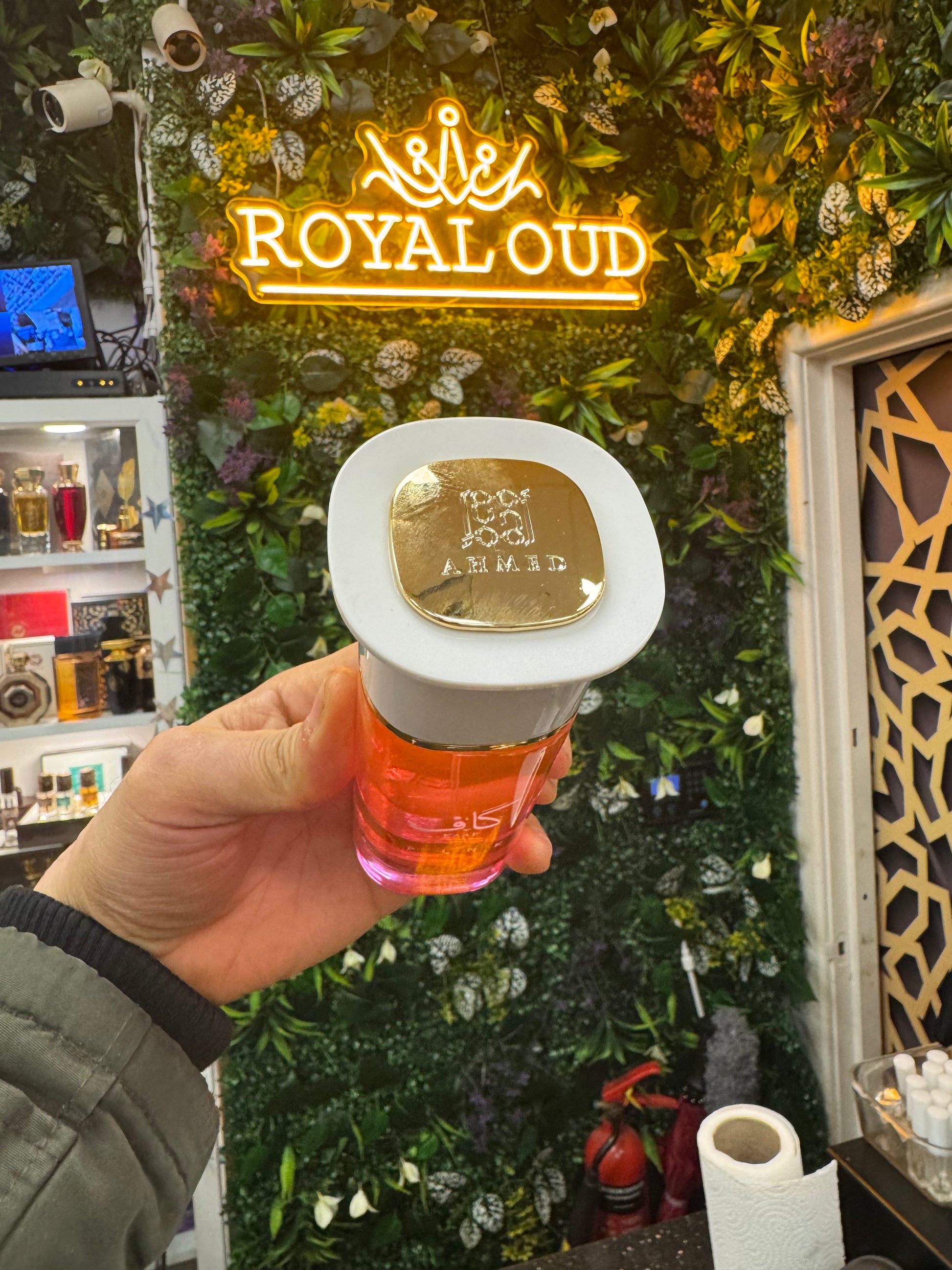 Hand holding a perfume bottle with a 'Royal Oud' neon sign in the background