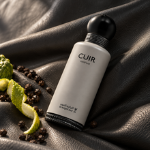 Cuir Parfum bottle lying diagonally on charcoal leather beside bergamot peel and black peppercorns